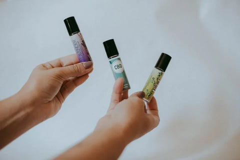 Hands holding three CBD essential oil roll-on bottles against a plain background.