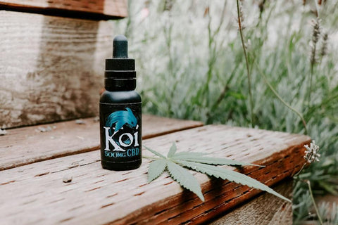 Koi 500mg CBD oil dropper bottle next to a cannabis leaf on a rustic wooden surface.